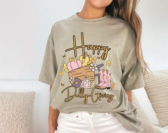 May include: A light olive green t-shirt with a graphic that says "Happy Dolly Giving." The graphic features a wooden cart with pumpkins, flowers, and cowboy boots. The design includes butterflies and fall leaves.