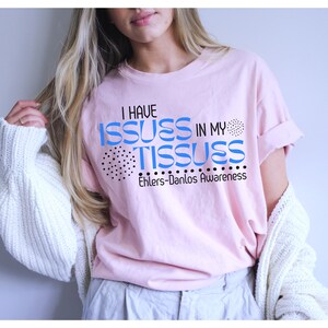 May include: A light pink t-shirt with the text "I HAVE ISSUES IN MY TISSUES" in blue, with black dot accents. The text "Ehlers-Danlos Awareness" is below the main text. The shirt is a casual style.