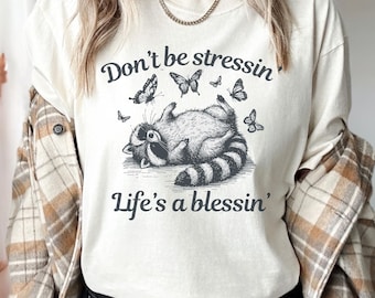 May include: A white t-shirt with a graphic of a raccoon laying on its back with butterflies flying around it. The text "Don't be stressin' Life's a blessin'" is printed on the shirt.