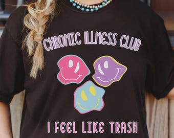 May include: Black t-shirt with the text "CHRONIC ILLNESS CLUB" and "I FEEL LIKE TRASH" in pink. The shirt features three smiley face graphics in pink, purple, and blue. The person is wearing a turquoise necklace and a decorative belt buckle.