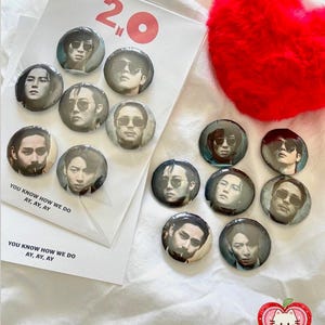 May include: A collection of round pinback buttons featuring images of people wearing sunglasses. Some buttons are arranged on a white card with the text "2.0" and "YOU KNOW HOW WE DO AY, AY, AY". A red heart-shaped plush is in the background.