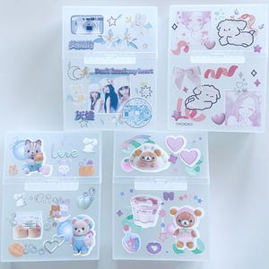 May include: Five clear plastic storage boxes decorated with stickers. The stickers feature images of cameras, cartoon characters, hearts, and text. The boxes are arranged in a vertical stack, showcasing a variety of designs and themes.