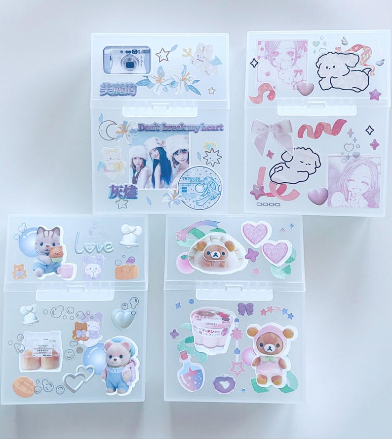 May include: Five clear plastic storage boxes decorated with stickers. The stickers feature images of cameras, cartoon characters, hearts, and text. The boxes are arranged in a vertical stack, showcasing a variety of designs and themes.