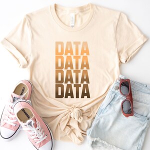 Puede incluir: Una camiseta beige con la palabra "DATA" repetida cuatro veces en un degradado de colores marrón y naranja. La camiseta se combina con pantalones cortos de mezclilla y zapatillas rosas.