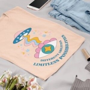 May include: A light pink t-shirt with a graphic print of a cartoon eye with a star inside, a flying saucer, and a rainbow. The text "LIMITLESS POSSIBILITIES DIFFERENT ABILITIES" is printed on the shirt.