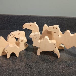 May include: A collection of wooden camel figurines in various sizes. The camels are light-colored, with simple, carved details. The set includes camels in standing and resting positions, suitable for children's play or decorative purposes.