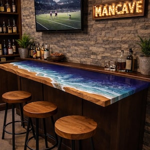 May include: A custom bar with a unique ocean-themed epoxy resin countertop. The bar features a wooden base, three wooden stools with black metal legs, and a "MANCAVE" sign. A TV displays a sports game.