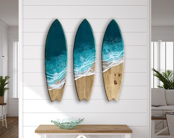 Op de afbeelding: Drie houten wanddecoraties in de vorm van surfplanken met een tealblauw en wit harsontwerp van een oceaan golf.