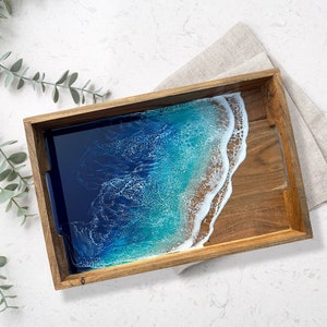 May include: Wooden serving tray with a resin ocean design. The tray features a rectangular shape with a dark wood frame. The ocean design incorporates shades of blue and white, resembling waves and a sandy shore. A linen napkin rests beside the tray.