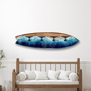 May include: A wooden surfboard with a resin coating depicting three whale tails breaking the surface of the ocean. The surfboard is mounted on a white wall above a wooden bench with white cushions.