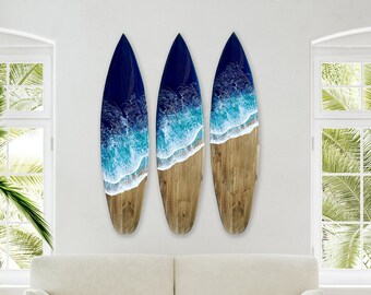 Op de afbeelding: Drie surfboard-vormige wandkunstwerken. Elk heeft een donkerblauwe bovenkant, een wit en turquoise golfontwerp en een lichtbruine houten basis. De kunst wordt tentoongesteld op een witte muur, waardoor een kustthema ontstaat.