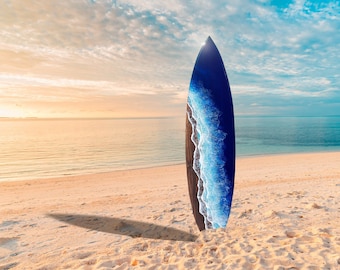 Op de afbeelding: Een blauw en zwart surfboard met een golfontwerp staat op een zandstrand met een heldere blauwe oceaan en een bewolkte lucht op de achtergrond.