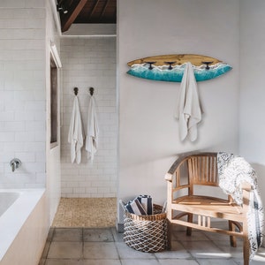 Peut inclure: Intérieur de salle de bain avec un porte-serviettes en forme de planche de surf, orné d'un motif de vagues bleues et de trois crochets. Des serviettes blanches sont suspendues au porte-serviettes et aux crochets muraux. Une chaise en bois et un panier tressé avec des serviettes sont également présents.