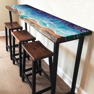 May include: A long, wooden table with a blue and green epoxy resin top that resembles an ocean wave. The table is supported by black metal legs and has three matching black metal stools with wooden seats.