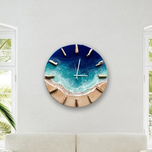 Puede incluir: Un reloj de pared redondo con una escena de playa. La esfera del reloj representa un océano azul con olas blancas rompiendo en una playa de arena. Las manecillas del reloj son plateadas.