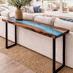 May include: A long, wooden console table with a blue resin river design. The table has a natural wood grain finish and black metal legs. A woven vase with greenery sits on the table, which is placed in front of a white sofa.