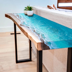 May include: A live edge console table with a resin top depicting an ocean scene. The table has a dark wood base and black metal legs. Two small potted plants sit on the table.
