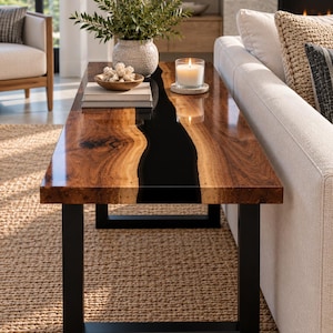 Pode incluir: Uma mesa de centro retangular com um tampo de madeira de borda natural e uma incrustação central de resina preta, apoiada por uma estrutura de metal preto. A superfície da mesa contém um vaso texturizado com plantas, uma tigela e uma vela.
