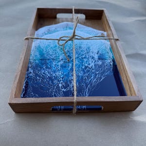 May include: Wooden serving tray with a rectangular shape and cut-out handles. The tray contains a set of coasters with a blue and white ocean wave design. The coasters are tied together with twine.