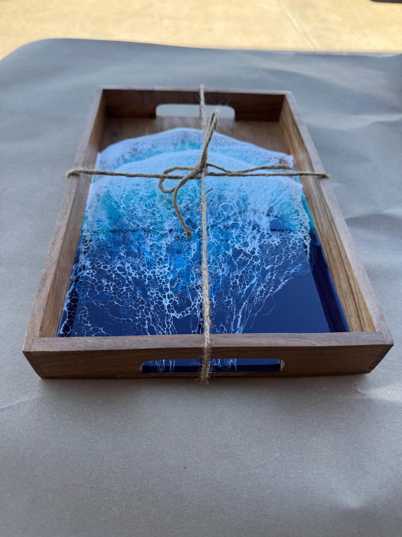 May include: Wooden serving tray with a rectangular shape and cut-out handles. The tray contains a set of coasters with a blue and white ocean wave design. The coasters are tied together with twine.