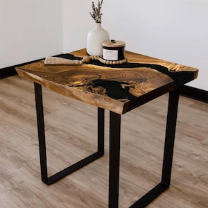 May include: A handcrafted side table with a live edge wooden top and black epoxy resin inlay. The table has a black metal base and is decorated with a white vase, candle, and wooden beads. The table measures approximately 24 inches long, 24 inches wide, and 20 inches high.