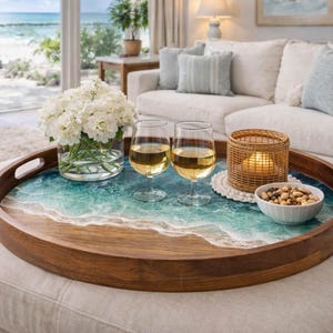 May include: A round wooden serving tray with a beach-themed resin design. The tray holds two wine glasses, a small bowl of nuts, a woven candle holder, and a vase of white flowers. The tray is on a white ottoman.
