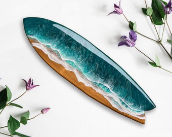 Op de afbeelding: Een houten serveerplank in de vorm van een surfplank met een harsontwerp van de oceaan. De plank heeft een bruine houten basis met een turquoise en wit golfpatroon. Paarse bloemen en groene bladeren zijn rond de plank gerangschikt.