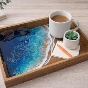 May include: A wooden serving tray with a resin ocean design. The tray holds a white mug of coffee, a small succulent in a white pot, a notepad, and a pen. Black glasses rest on the tray.