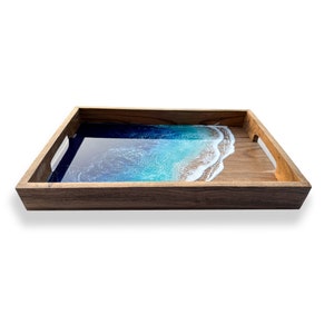 May include: Wooden serving tray with a resin ocean design. The tray features a dark brown wooden frame and handles. The interior has a blue and white resin design that resembles a beach scene.