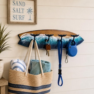 Puede incluir: Un perchero de pared con forma de tabla de surf de madera con ganchos de cola de ballena, con una escena oceánica pintada. Una bolsa de playa, toallas, llaves y una correa para perros cuelgan de los ganchos. Un letrero de madera arriba dice "SAND SALT SURF".