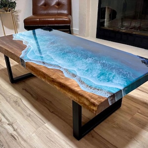 Peut inclure: Table basse rectangulaire avec une base en bois à bord brut et des pieds en métal noir. Le plateau présente un motif en résine représentant des vagues océaniques bleues et blanches, créant un thème côtier.
