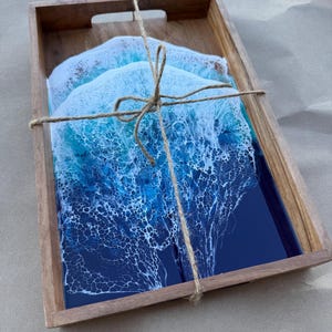 May include: A wooden serving tray with three coasters. The coasters have a blue and white ocean wave design. The tray and coasters are tied together with twine. The coasters have a glossy finish.