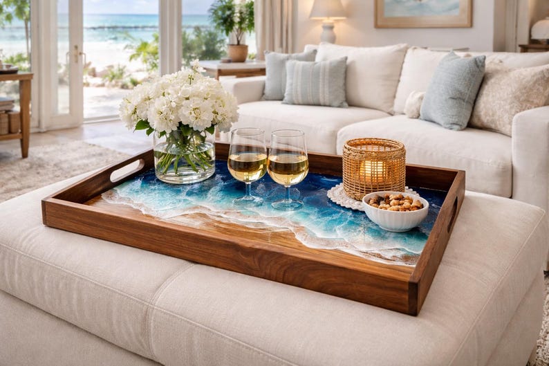 May include: A wooden serving tray with a resin ocean design, holding two wine glasses, a candle in a woven holder, a bowl of nuts, and a vase of white flowers. The tray sits on a white ottoman in a living room.