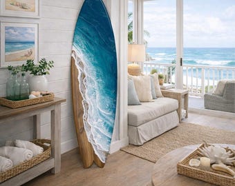 Op de afbeelding: Een decoratief surfboard met een blauw en wit golfontwerp, leunend tegen een witte muur. Het surfboard heeft een houten basis en wordt tentoongesteld in een woonkamer met kustthema en uitzicht op de oceaan.
