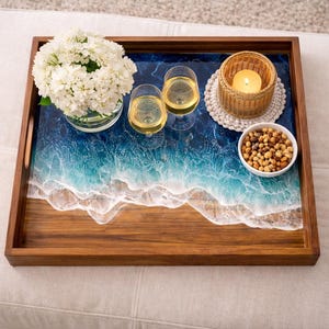 May include: A wooden serving tray with a beach-themed resin design. The tray holds a bouquet of white flowers, two glasses of wine, a candle, and a bowl of snacks. The tray is brown with a blue and white wave design.