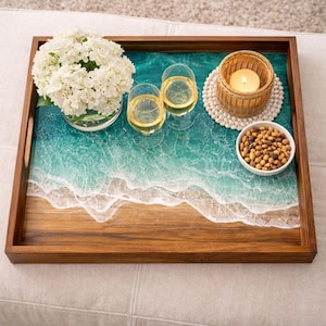 May include: A wooden serving tray with a resin ocean design, holding two wine glasses, a candle, a bowl of snacks, and a vase of white flowers. The tray is brown with a turquoise and white wave pattern.