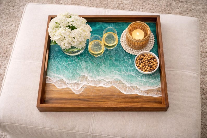 May include: A wooden serving tray with a resin ocean design, holding two wine glasses, a candle, a bowl of snacks, and a vase of white flowers. The tray is brown with a turquoise and white wave pattern.