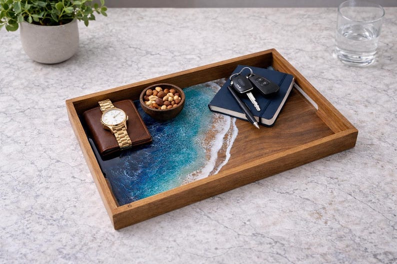 May include: A rectangular wooden tray with a resin design depicting a beach scene. The tray holds a gold watch, a brown wallet, a small wooden bowl of nuts, a blue notebook, keys, and a pen. The tray is on a light gray surface.