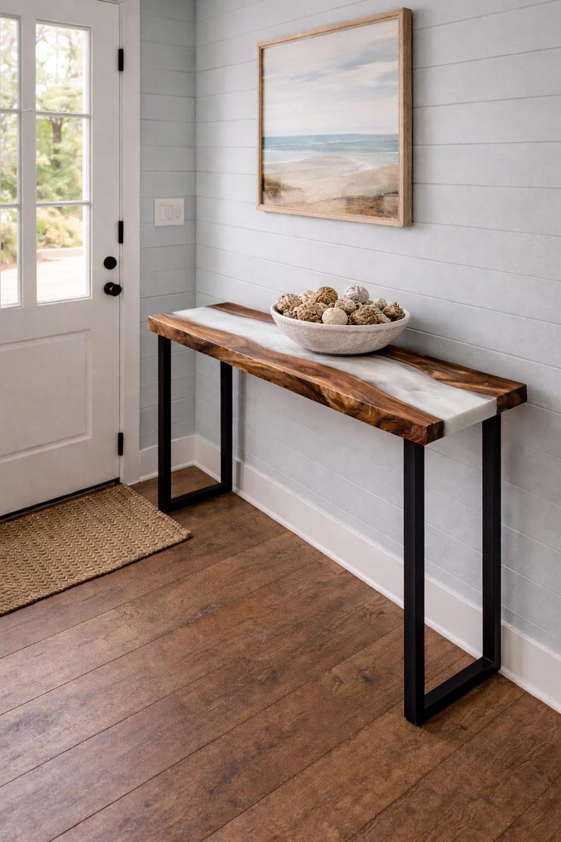 May include: A wooden console table with a live edge and a white resin river. The table has black metal legs and a bowl filled with decorative spheres. A framed beach scene hangs on the wall above the table.