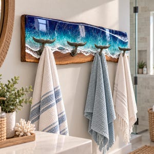 Könnte beinhalten: Ein wandmontierter Holzkleiderhaken mit Naturkante und einem blauen sowie weißen Harz-Ozeanwelleneffekt. Die Leiste verfügt über vier Metallhaken in Form von Walflossen, an denen jeweils ein weißes, blaues oder cremefarbenes Handtuch hängt.