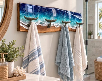 May include: A wall-mounted wooden coat rack featuring a live edge design with a blue and white resin ocean wave effect. The rack includes four metal hooks shaped like whale tails, each holding a hanging towel in white, blue, or cream.