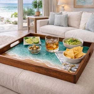 May include: A wooden serving tray with a beach-themed design, featuring a turquoise and white resin ocean scene. The tray holds a glass of whiskey, guacamole, chips, olives, and lime slices, perfect for entertaining.