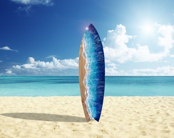 Op de afbeelding: Een surfboard met een uniek ontwerp staat op een zandstrand. Het board heeft een houten basis met een blauw en wit golfachtig ontwerp op de bovenste helft. De achtergrond toont een helderblauwe lucht, oceaan en witte wolken.