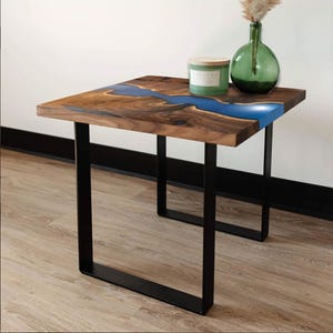May include: A square wooden side table with a live edge design and a blue resin river. The tabletop is supported by black metal legs. A green candle and a green glass vase with dried flowers are on the table.