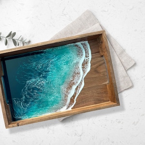 May include: Wooden serving tray with a resin ocean design. The tray features a rectangular shape with a natural wood frame. The resin art depicts a turquoise ocean with white foamy waves. A linen napkin rests beside the tray, and a sprig of greenery is in the corner.