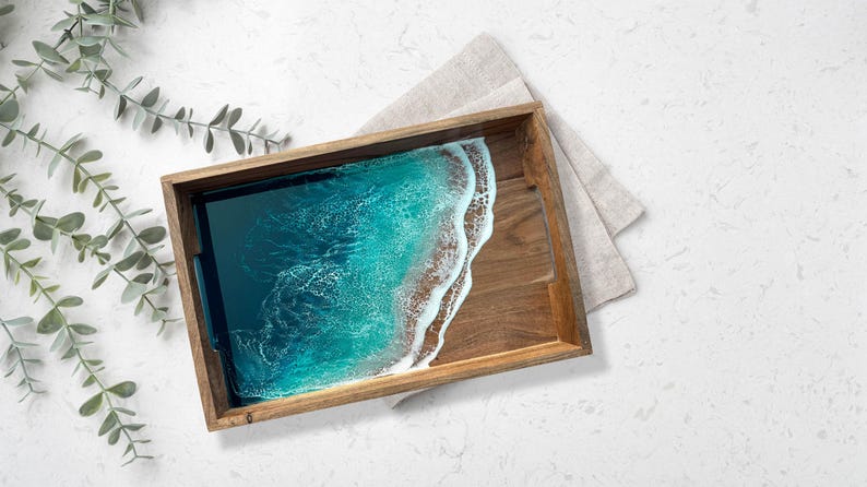 May include: Wooden serving tray with a resin ocean design. The tray features a rectangular shape with a natural wood frame. The resin art depicts a turquoise ocean with white foamy waves. A linen napkin rests beside the tray, and a sprig of greenery is in the corner.