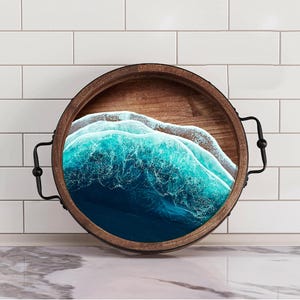 May include: A round wooden serving tray with black metal handles. The tray features a resin inlay of an ocean wave with blue and white colors.