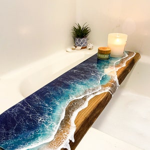 May include: A wooden bath tray with a resin ocean wave design. The tray has a natural wood finish and features a white wave crashing on a blue ocean background. The tray is decorated with a small potted plant, a candle, and a jar labelled "Releaf Balm".