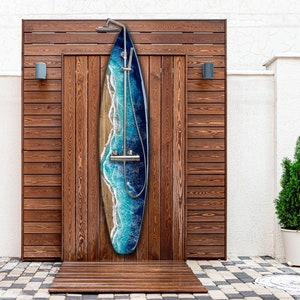 May include: An outdoor shower with a surfboard design. The shower has a wooden frame and a surfboard-shaped panel with a blue and white wave design. The shower head and fixtures are metal. The shower is set on a wooden floor.