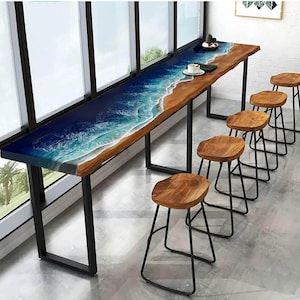 May include: A long wooden table with a blue and white epoxy resin top featuring a wave design. The table is supported by black metal legs and is surrounded by four wooden bar stools with black metal frames.